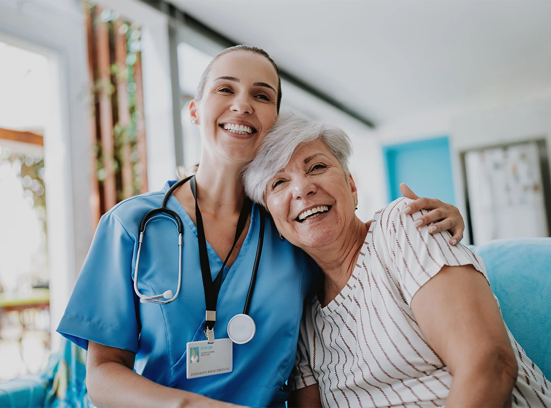 Smiling nurse from our agency embraces a cheerful senior patient in a bright, welcoming care facility.
