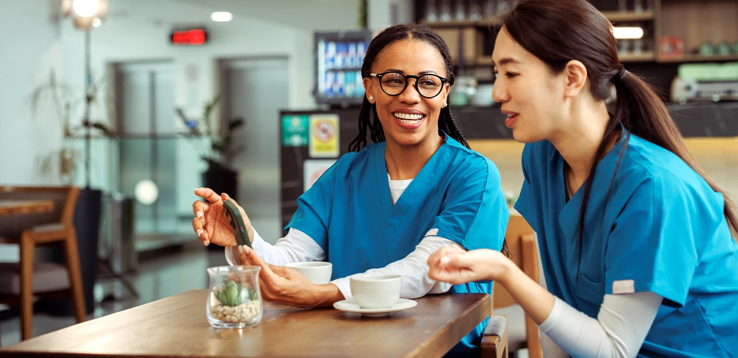 2 nurses relaxing and smiling over coffee during a break, reflecting a welcoming and approachable healthcare environment.